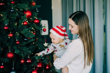 Çocuğun ilk Noel 'i. Anne, parlak bir Noel ağacı olan karanlık bir arka planda, Noel Baba şapkalı küçük bir çocuğu kucağında tutuyor. Noel konsepti.