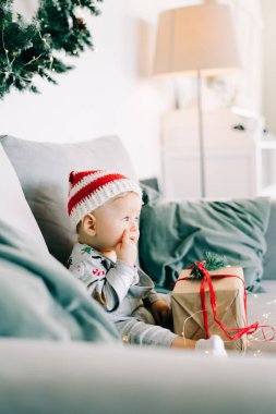 Düşünceli küçük çocuk Noel Baba şapkası ve Noel kostümü içinde gri koltukta oturuyor. Kırmızı kurdeleli büyük bir hediye, beyaz duvara karşı doğal köknar çelengi. Noel konsepti..