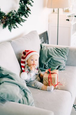 Düşünceli küçük çocuk Noel Baba şapkası ve Noel kostümü içinde gri koltukta oturuyor. Kırmızı kurdeleli büyük bir hediye, beyaz duvara karşı doğal köknar çelengi. Noel konsepti..