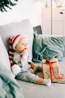Düşünceli küçük çocuk Noel Baba şapkası ve Noel kostümü içinde gri koltukta oturuyor. Kırmızı kurdeleli büyük bir hediye, beyaz duvara karşı doğal köknar çelengi. Noel konsepti..