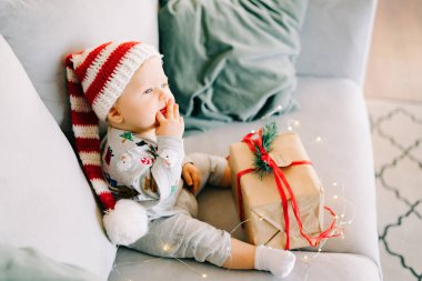 Düşünceli küçük çocuk Noel Baba şapkası ve Noel kostümü içinde gri koltukta oturuyor. Kırmızı kurdeleli büyük bir hediye, beyaz duvara karşı doğal köknar çelengi. Noel konsepti..