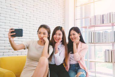 Güzel genç iş kadınları, arkadaşıyla cep telefonuyla ya da akıllı telefonuyla oturma odasında selfie çekerler. Mutlu arkadaşlık ilişkisi konsepti.