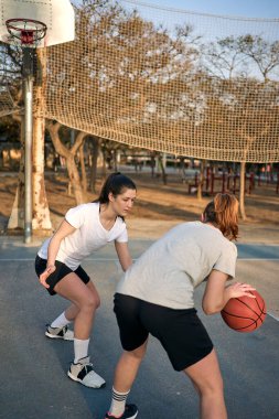 İki kız, birinin savunduğu, diğerinin güneşli bir sabahta saldırdığı şehir sahasında basketbol oynuyor..