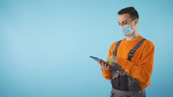 Mechanik z maską twarzy pracujący na tablecie odizolowanym w studio niebieskim tle — Wideo stockowe