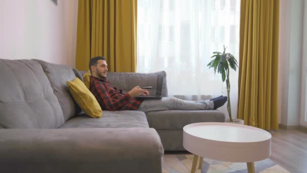 Man in square shirt Berbaring di sofa dengan laptop dan menyalakan TV menggunakan remote — Stok Video