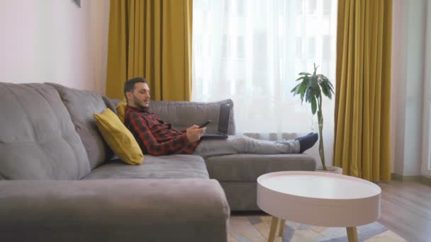 Jongeman werkt thuis bank met behulp van laptop en reiken naar de tv-afstandsbediening — Stockvideo