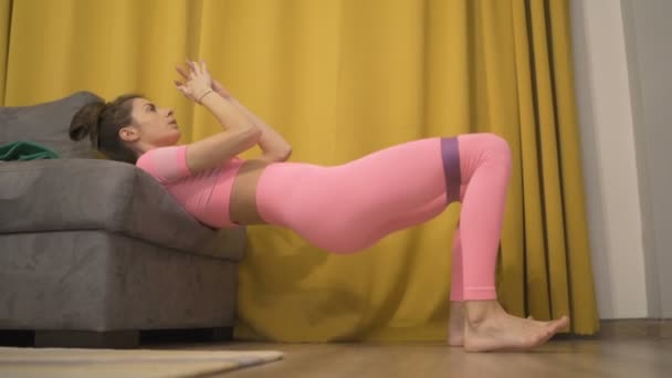 Autentisk video fitness flicka i rosa träning med gummi på soffan — Stockvideo