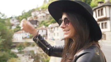 Siyah şapkalı, güneş gözlüklü, arka planda eski evleri olan bir gezgin kız.