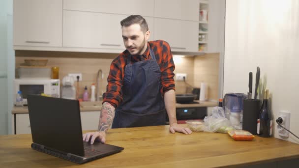 Önlüklü genç adam ev mutfağında dizüstü bilgisayarla çalışıyor. — Stok video