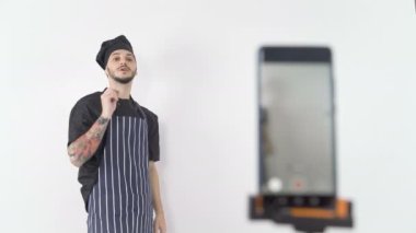 Önlüklü ve şapkalı gülümseyen profesyonel erkek aşçı akıllı telefondan internet üzerinden ders yayınladı.