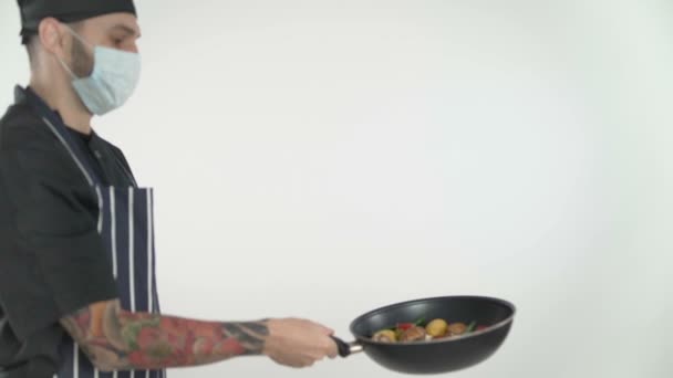 カメラの前で鍋に野菜を投げ顔マスクを持つ男性シェフ — ストック動画
