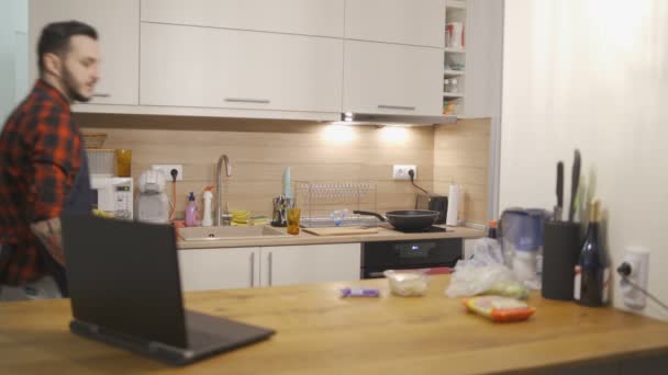 Önlüklü genç adam mutfak tezgahından ürünleri taşıyor ve dizüstü bilgisayarını kontrol ediyor. — Stok video
