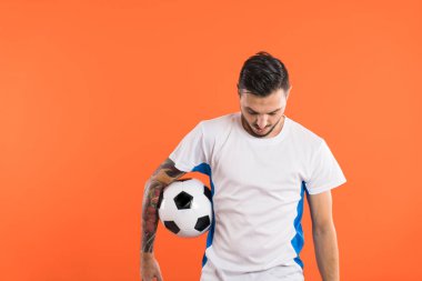 Futbolcu aşağıya bakıyor ve bir kolu turuncu arka planda izole edilmiş bir topu tutuyor.