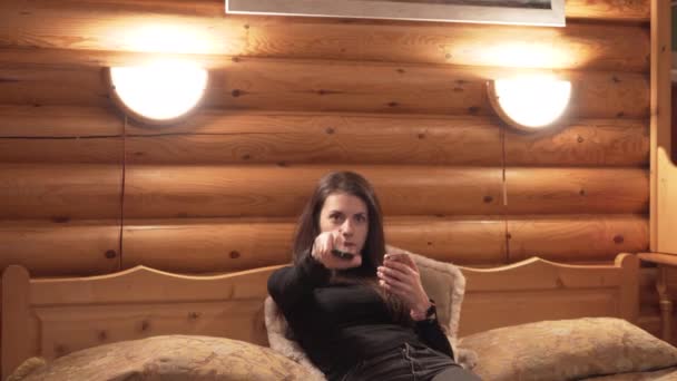Front view of woman switching TV channels from big bed in winter wooden cabin — Stock Video
