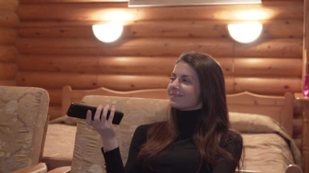 Woman with a tv remote control sitting in a winter wooden cabin — Stock Video