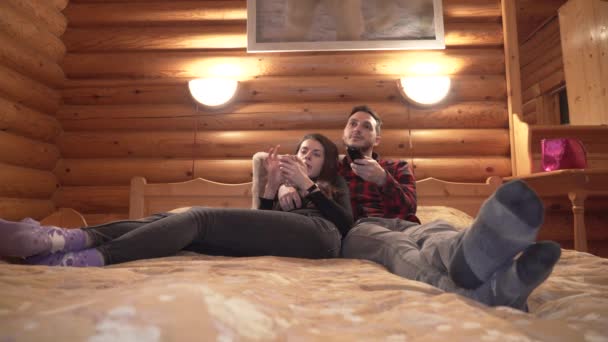 Young couple lying in bed in log cabin. Woman using smartphone and man switching TV channels with remote — Stock Video