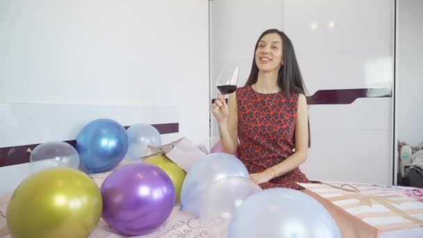 Krásná narozeninová žena s červenými šaty sedí na posteli s balónky a drží sklenici červeného vína — Stock video