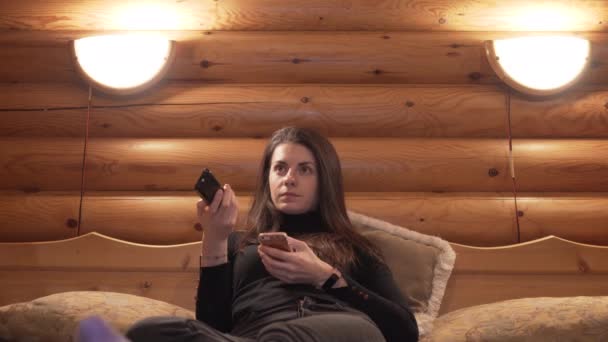 Mulher jovem trocando canais de TV de cama grande na cabine de madeira de inverno — Vídeo de Stock
