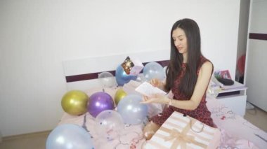 Güzel bir kadın renkli balonlarla yatakta oturuyor ve konfeti bombası doğum günü kartını açıyor.