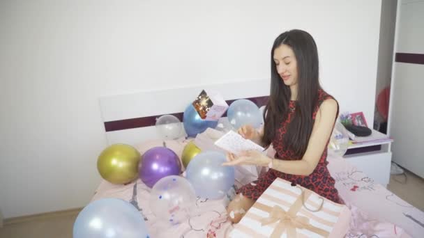 Gyönyörű nő ül az ágyon színes lufikkal és konfetti bomba szülinapi kártyát nyit — Stock videók