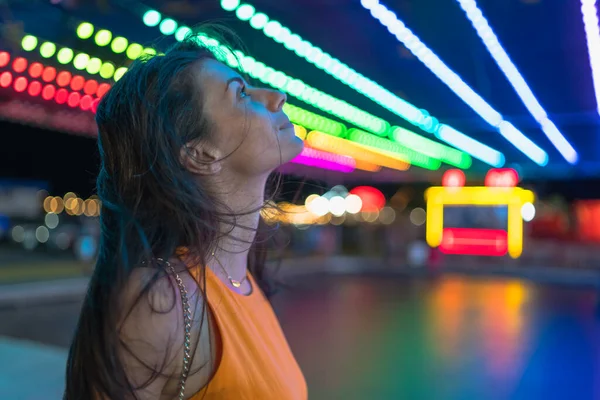 Foto lateral de chica reflexiva con el pelo soplado mirando las luces en el parque de atracciones por la noche Imágenes De Stock Sin Royalties Gratis