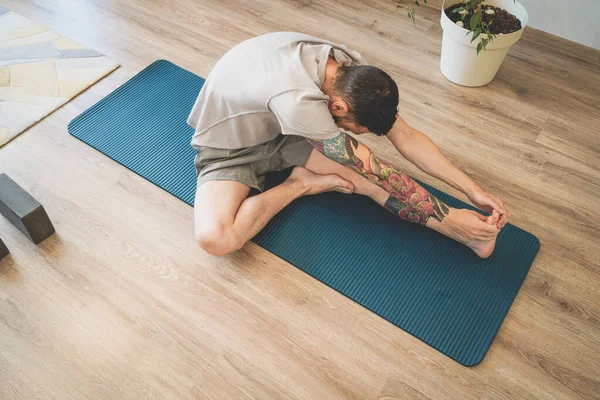 Vista superior del joven tatuado estirándose sobre una esterilla de yoga alcanzando dedos de los pies con las manos Imagen De Stock