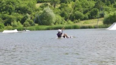 Çekme halatıyla çekilen genç kadın kalkıyor ve rampaların yanında wakeboard sürüyor.