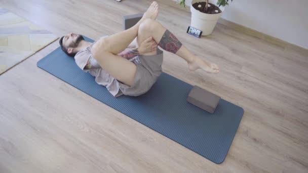 Young man practicing Pavanamuktasana yoga indoors at home — Stock Video