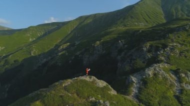 Dağın tepesindeki kayalıklarda oturan, elinde harita tutan yalnız yürüyüşçünün insansız hava aracı görüntüsü.