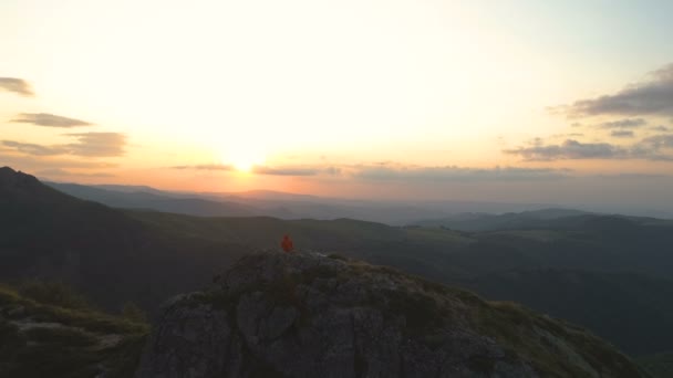 Wandelaar in oranje jasje rust op rotsachtige bergrand kijkend naar de zonsondergang — Stockvideo