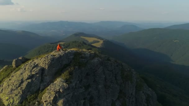 Mężczyzna wędrowiec siedzi na szczycie skalistego szczytu góry, patrząc w dół głębokiej doliny — Wideo stockowe