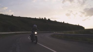 Gün batımında virajlı yolda motosiklet süren bir adam.