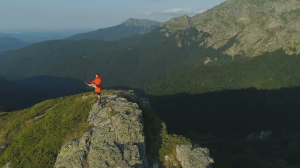 Wędrowiec w pomarańczowej kurtce stojący na szczycie góry i trzymający mapę, rozglądający się dookoła. — Wideo stockowe
