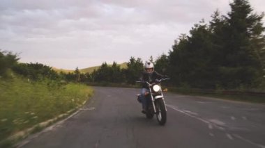 Yeşil tepeli virajlı yolda motosiklet süren bir adam.