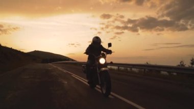 Yavaş çekimde gün batımına karşı dağ yolunda motosiklet süren tek sürücünün silueti.