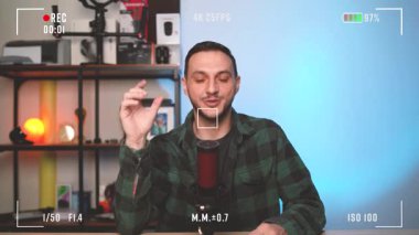 Genç bir adam, evinde bir podcast sunarken stüdyo mikrofonuna açık bir şekilde konuşuyor. Kendine güvenen ve profesyonel sunumuyla izleyicileri kendine çekiyor..