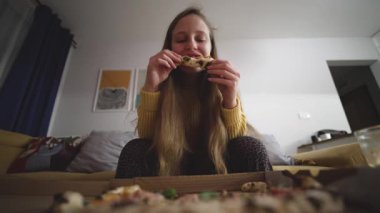 Ön koltukta beyaz kadın pizza diliminin tadını çıkarıyor, sıcak bir oturma odasında pizza kutusuyla ön planda, uzun sarışın.