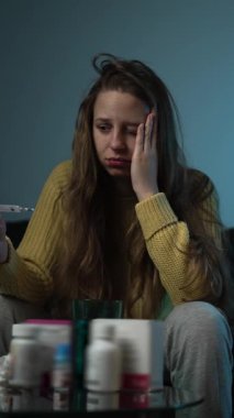 Genç beyaz kadın, burnunu mendile sümkürürken koltukta hasta yatıyor, etrafı ilaç şişeleriyle çevrili ve kullanılmış.