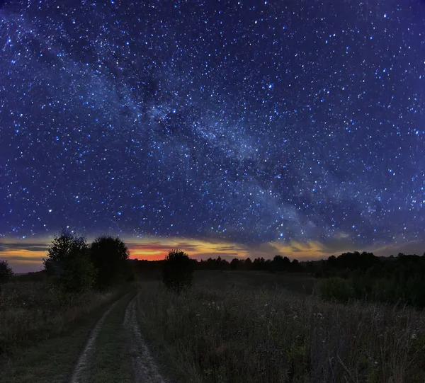 Starry summer night landscape - Milky Way over country road on meadow. Night view of natural glowing stars on dark blue sky. Narrow strip of bright sunset light is on horizon