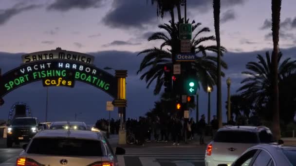 遅い動きのカメラで夕暮れ時にサンタモニカ桟橋の入り口前方に移動し、観光客が車を移動歩いてください。カリフォルニア州サンタモニカ- 2020年1月 — ストック動画