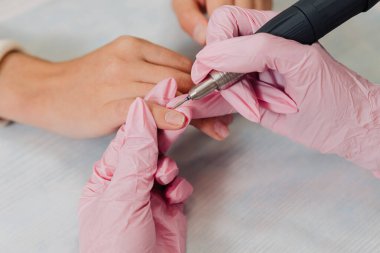 Manikür işlemi. Pembe lastik eldivenli bir usta elektrikli tırnak törpüsüyle tırnaklarını parlatır. Kadın elleri yakın..