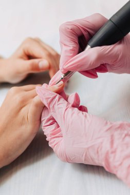 Manikür işlemi. Pembe lastik eldivenli bir usta elektrikli tırnak törpüsüyle tırnaklarını parlatır. Kadın elleri yakın..