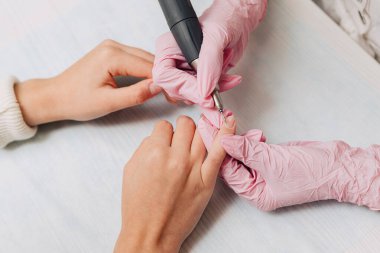 Manikür işlemi. Pembe lastik eldivenli bir usta elektrikli tırnak törpüsüyle tırnaklarını parlatır. Kadın elleri yakın..