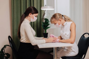 Manikür işlemi. Evde çalış. Efendi manikür yapıyor. Tıbbi maskeli iki kadın..