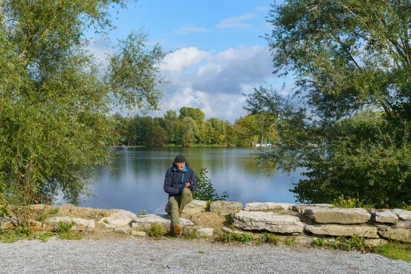 男がスマートフォンで湖の岸に石の上に座っている 白い雲と緑の木と青空 — ストック写真