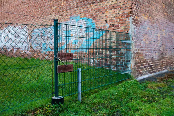 Chain link fence. Rusty riveted plates and padlock. There is a dirty brick wall behind the fence.