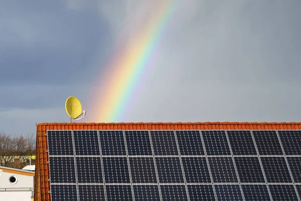 Rainbow over a tiled roof with solar panels. Satellite dish with a smile on the background of a dark sky.