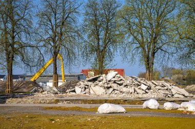 Yıkılmış bir binanın beton enkazı ve sarı bir kazıcı oku. Ön planda beyaz çantalar.
