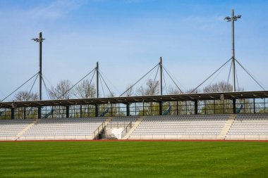Bir futbol stadyumunun boş tribünleri ve yeşil bir çimen. Stadyumun üstünde direkler var..