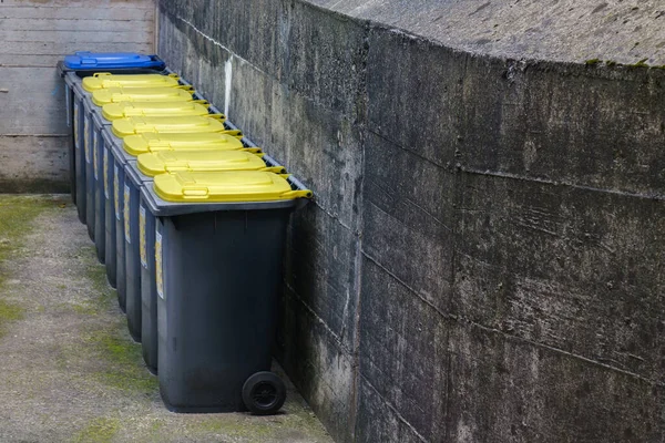 Sarı kapaklı çöp bidonları beton duvar boyunca sıralanmış. Mavi kapaklı son kutu..